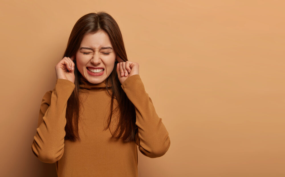 Distressed frustrated woman plugs ears, clenches teeth, hears bothering noise, avoids unpleasant sound, wears brown clothes, cannot hear anymore, listens friends playing, isolated on beige wall