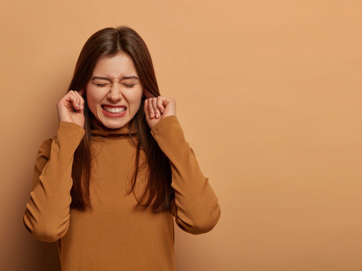 Distressed frustrated woman plugs ears, clenches teeth, hears bothering noise, avoids unpleasant sound, wears brown clothes, cannot hear anymore, listens friends playing, isolated on beige wall